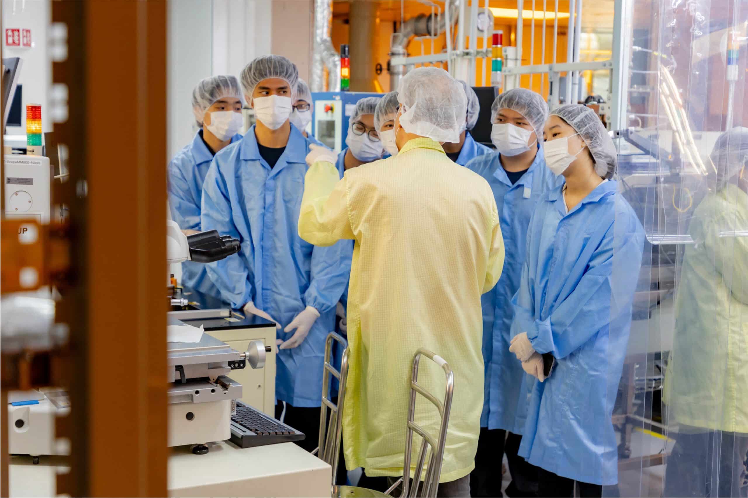 Engineer briefing trainees in cleanroom suits inside a semiconductor fabrication lab.